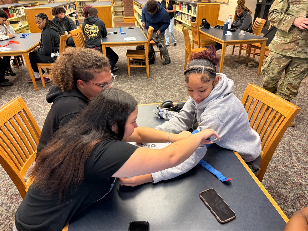 Three students are engaging in a Stop the Bleed Training in a school library. Other people can be seen in the background.