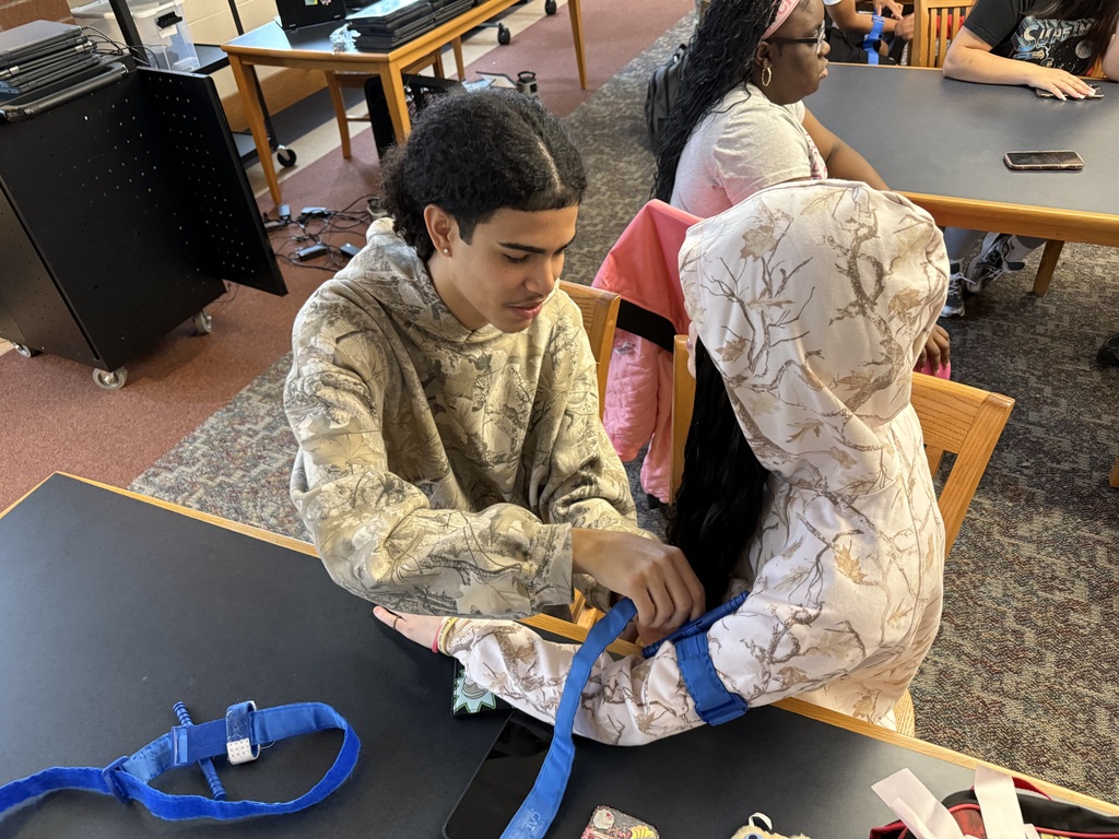 Two students are engaging in a Stop the Bleed Training in a school library. Other people can be seen in the background.