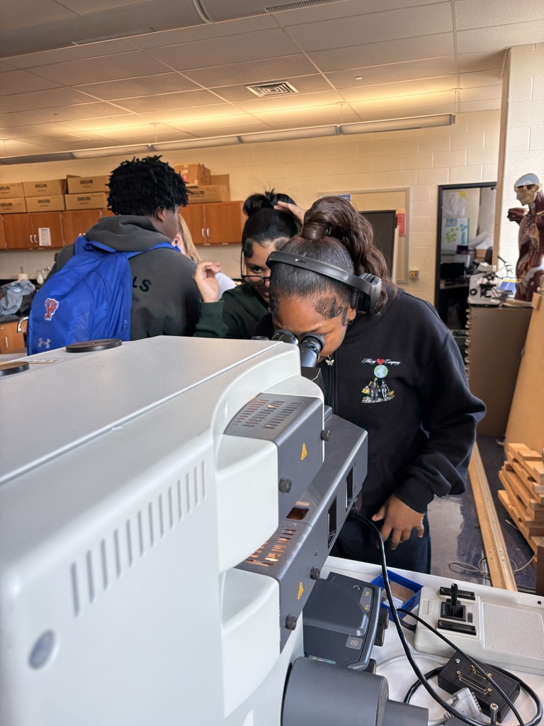 A student looking into a microscope in a classroom setting. Other people can be seen in the background.
