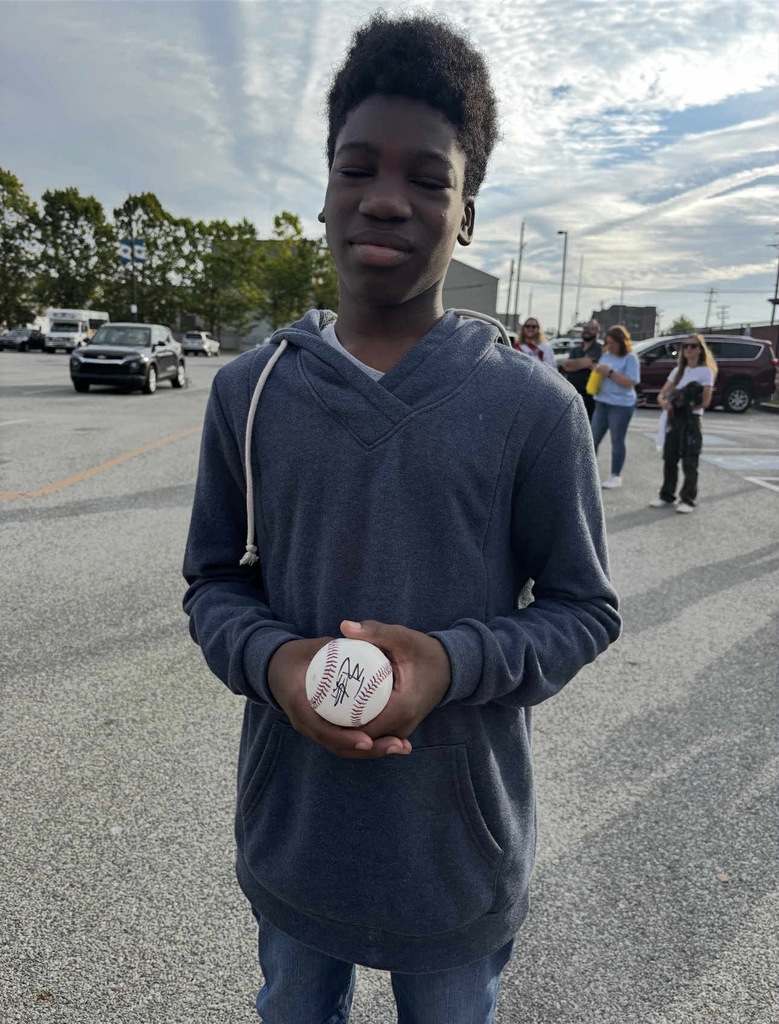 A student holding a baseball outside in a parking lot. Other people can be seen nearby. 