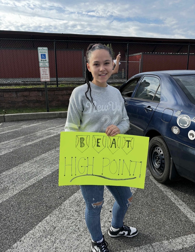 A student holding up a poster that says "Beat High Point!" outside in a parking lot. The student is standing near a car. 
