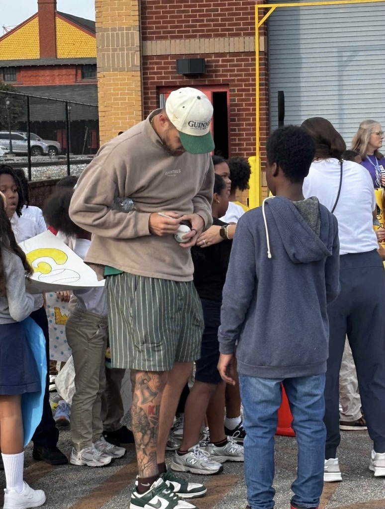 A man signing a baseball outside in a parking lot. Other people can be seen nearby.
