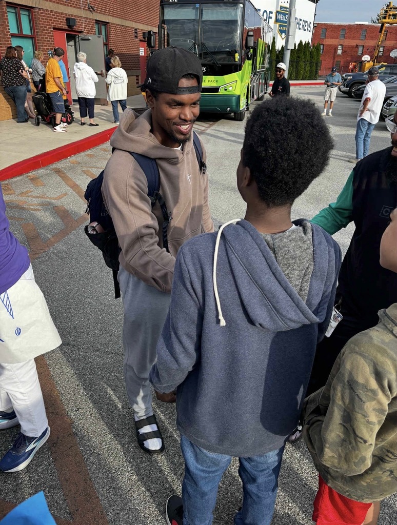 A man smiling and shaking a student's hand outside in a parking lot. Other people and a bus can be seen nearby.