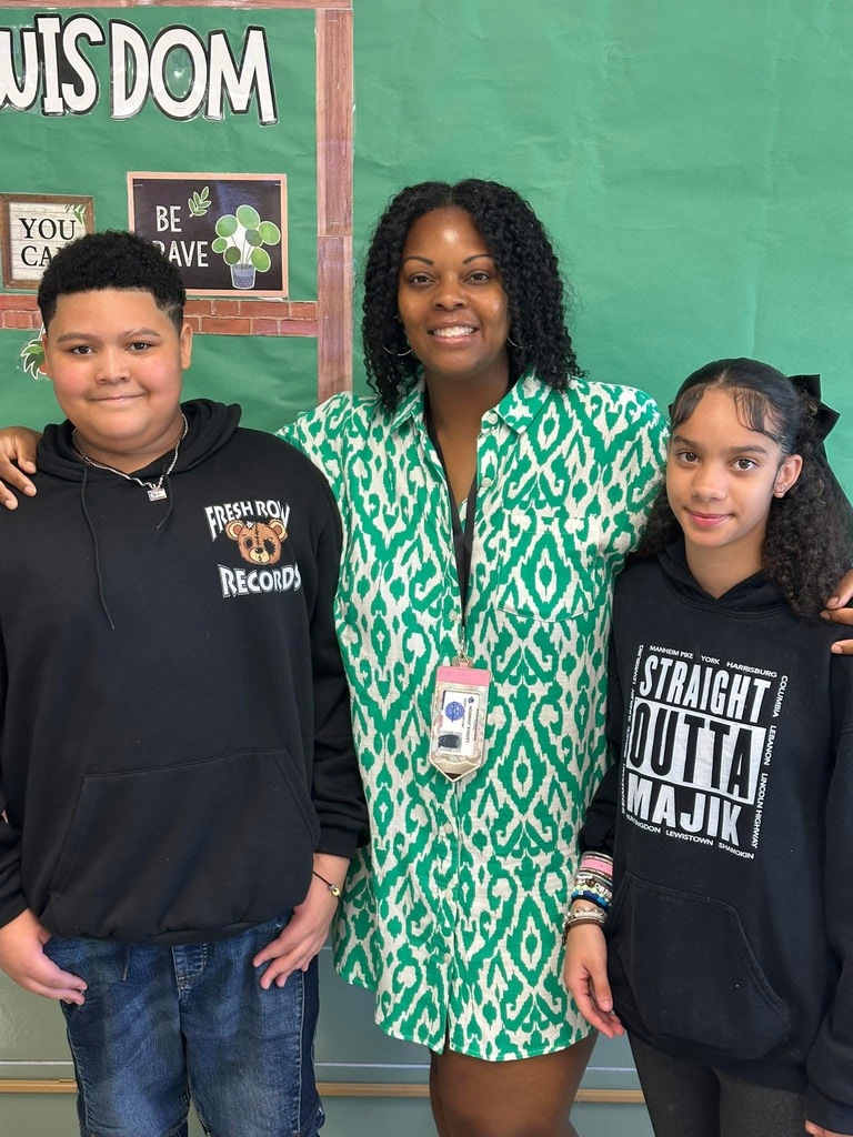 A district staff member and two students standing together and smiling in front of a bulletin board.