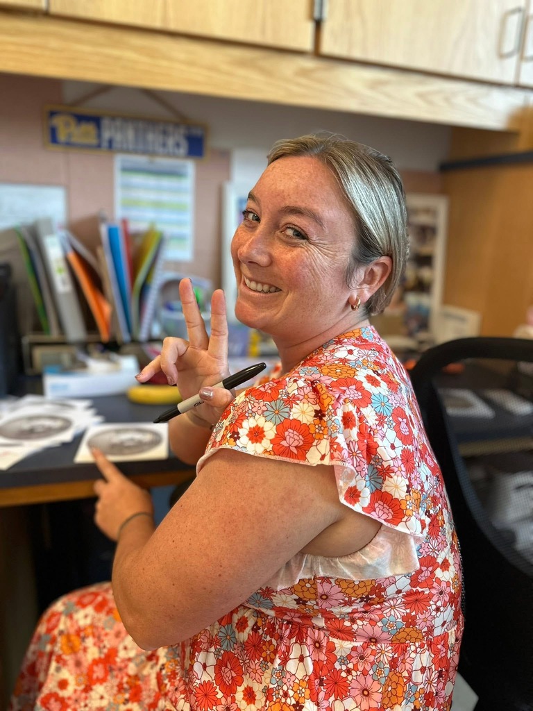 A district staff member smiling while sitting at a desk and holding up the peace sign. 