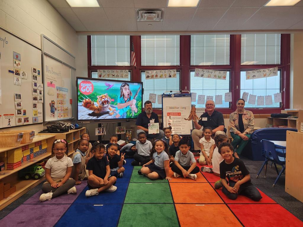 A diverse group of cheerful students are sitting on a colorful carpet in a classroom. Behind them are three adults and a display board showing educational content.