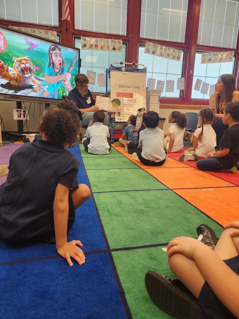 An adult is reading to a group of students who are sitting on a colorful carpet in a classroom.