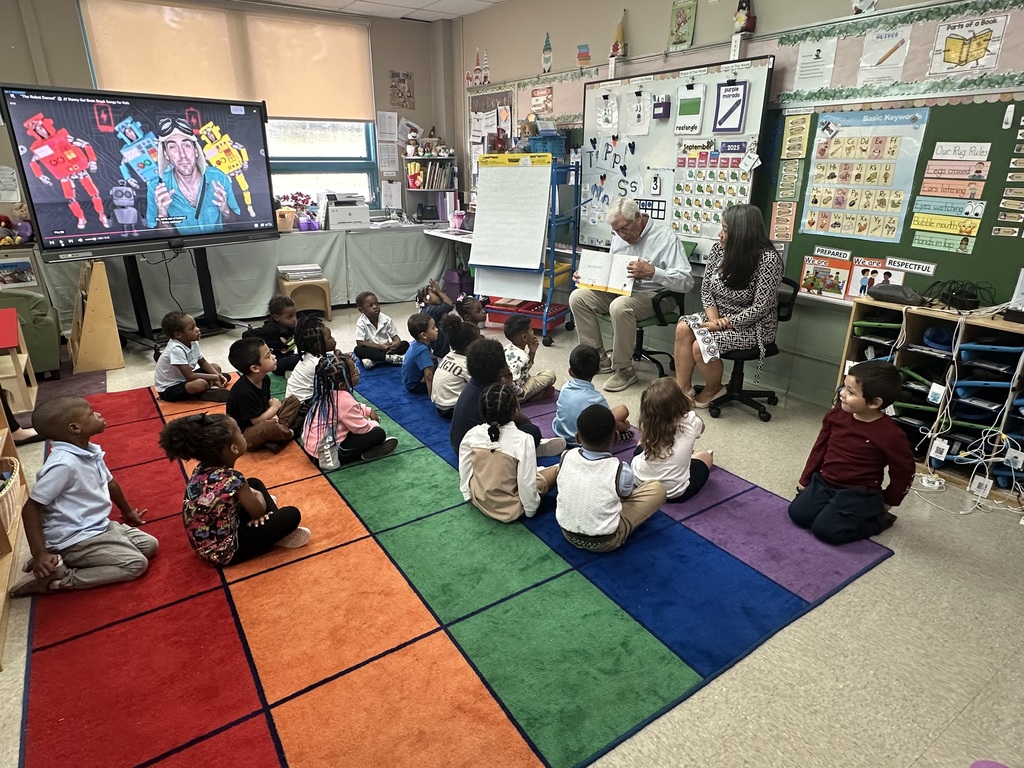 Two adults are reading to a group of students who are sitting on a colorful carpet in a classroom.