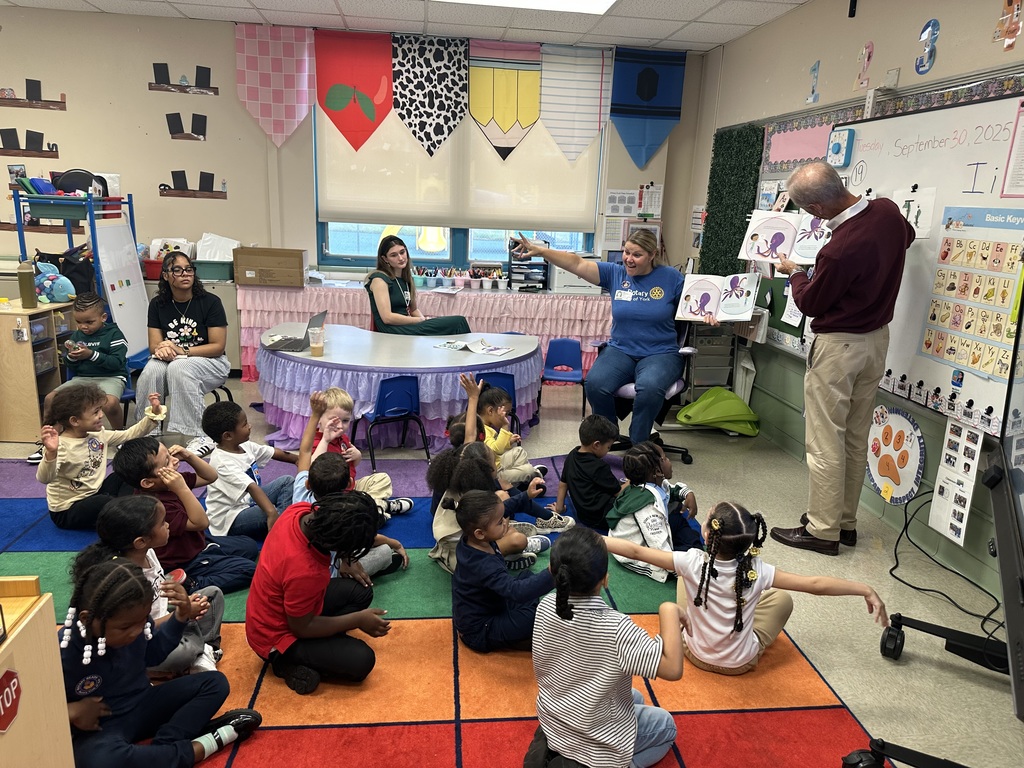 Two adults are reading to a group of students in a classroom. Two district staff members can be seen nearby sitting at a table.