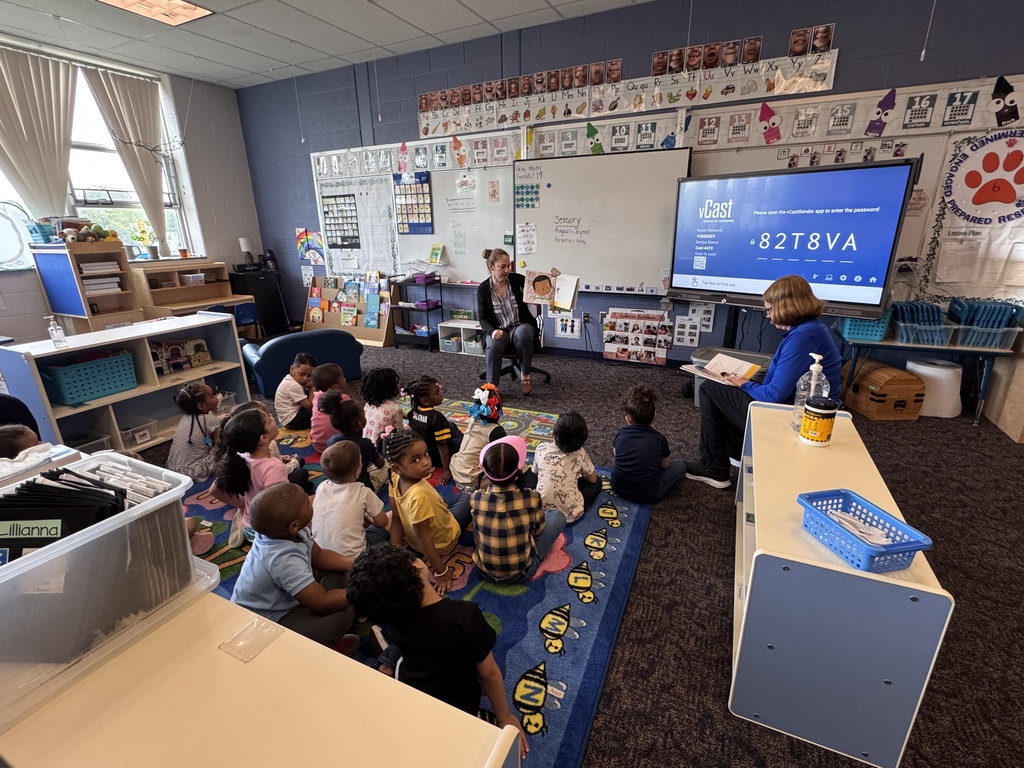 Two adults are reading to a group of students who are sitting on a colorful carpet in a classroom.