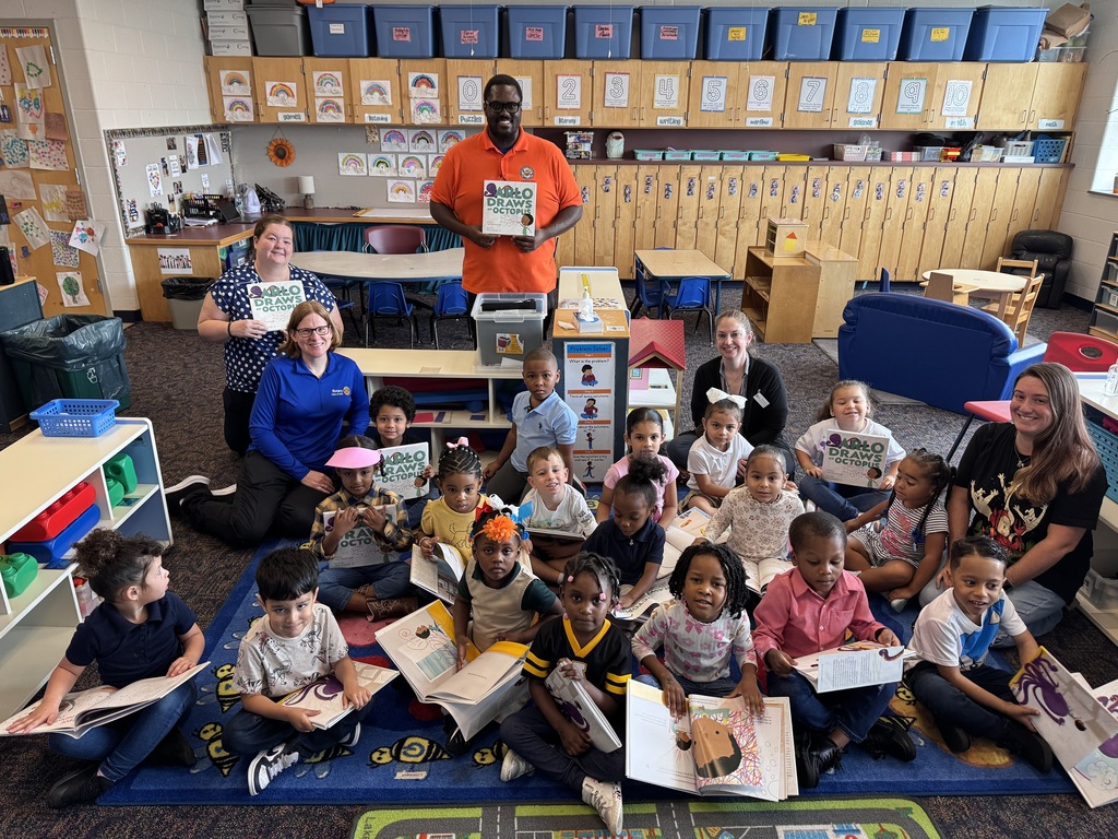 District staff members, a group of adults and a group of students are sitting and standing in a classroom. Some of the adults and students are holding books in their hands.