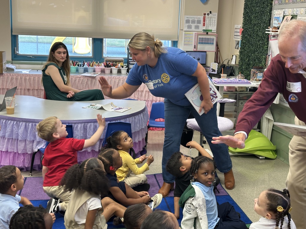 Two adults are giving high five's to a group students who are sitting on a carpet in a classroom. A district staff member can be seen sitting at a table nearby.