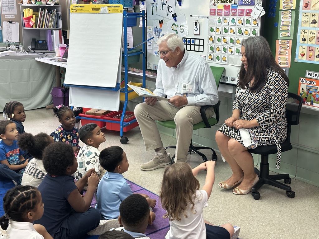 Two adults are reading to a group of students who are sitting on a colorful carpet in a classroom.