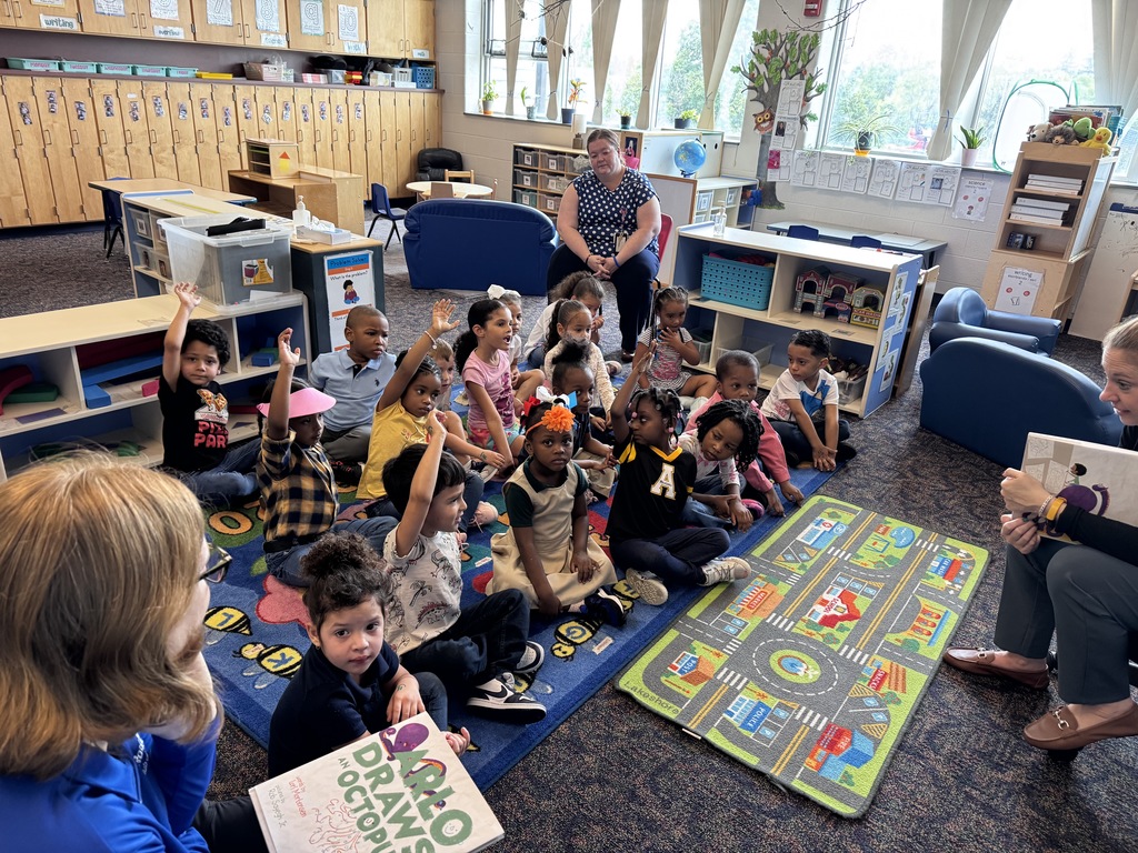 Two adults are reading to a group of students who are sitting on a colorful carpet in a classroom. A district staff member can be seen sitting in a chair near the students.
