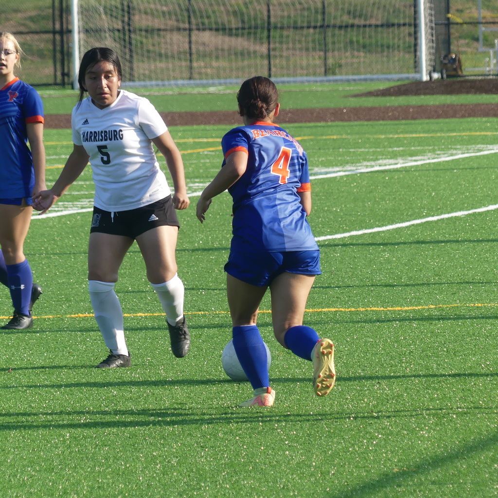York High Girls Soccer player running and kicking a soccer ball outside on a field. Another York High Girls Soccer player and a soccer player from the other team can be seen looking at the soccer ball.