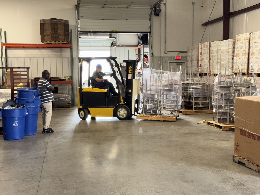 A district staff member is driving a yellow forklift and moving a pallet of stacked chairs in a warehouse. A man is standing nearby watching.