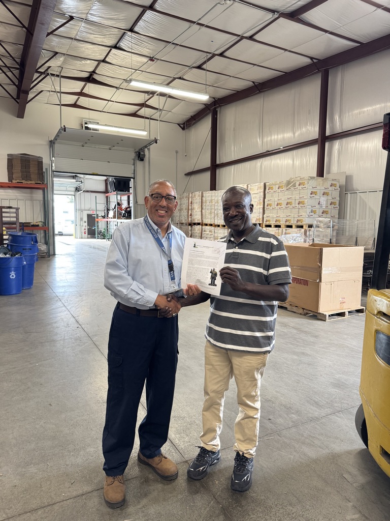 The School District of the City of York's Buildings and Grounds Supervisor, Mr. Muldrow standing in a warehouse with a man who is holding a certificate. Both men are smiling and shaking hands. Boxes and barrels can be seen in the background.