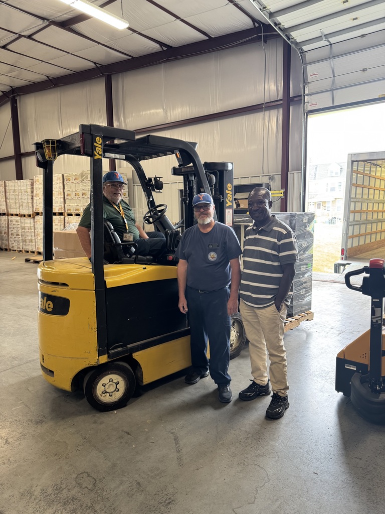 Two district staff members and a man are posing for a photo in a warehouse. One of the men is sitting on a yellow forklift with the other two men are standing beside him.