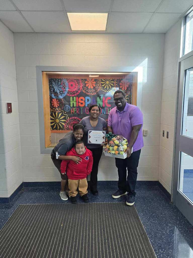 Ferguson PreK-8 Assistant Principal, Dr. Jones, a district staff member, a district parent and a student posing in front of a Hispanic Heritage Month banner. Dr. Jones is holding a fruit basket and the district parent is holding a certificate. 