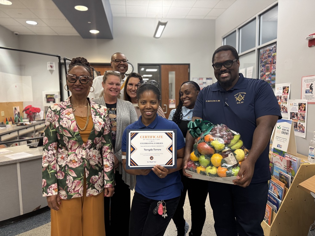 District leadership posing together in a lobby area. A district staff member is holding a certificate while another district staff member is holding a fruit basket.