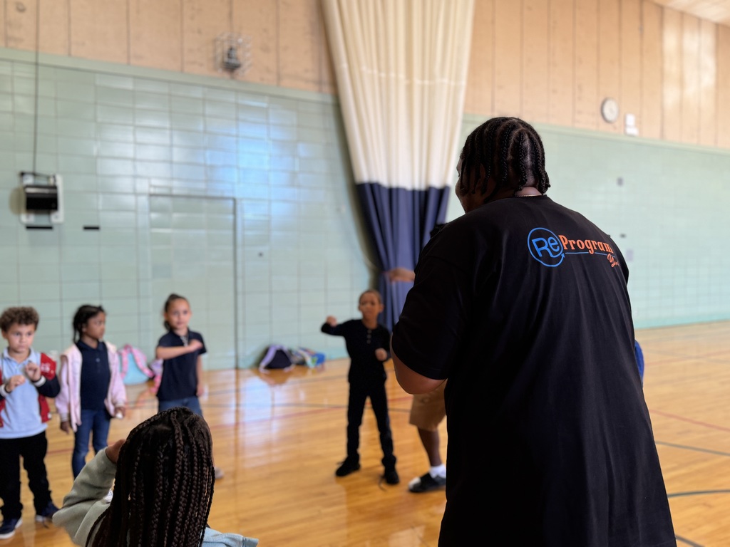 An adult wearing a "ReProgram" shirt is leading a group of young children in a school gym. The children are following the adult's instructions.