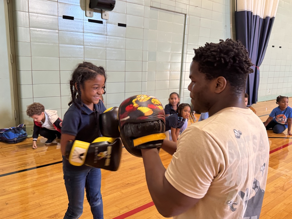 A young girl is practicing boxing with an adult in a school gym. The adult and the young girl are both wearing yellow boxing gloves. Other young children can be seen in the background.