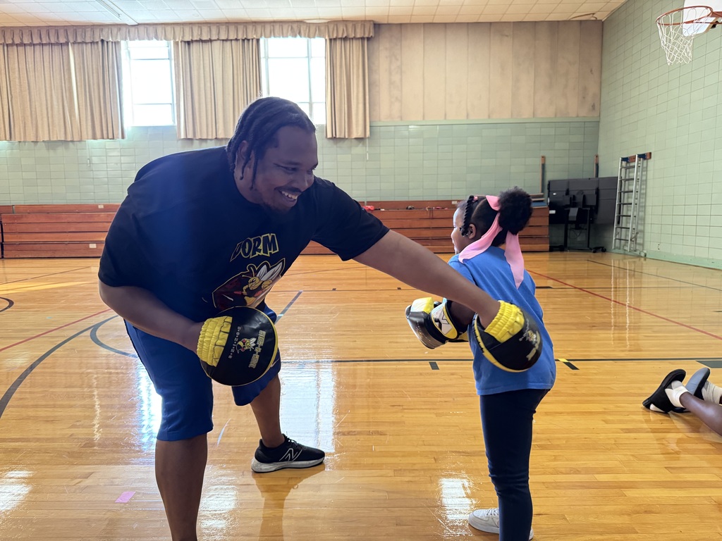 A young girl is practicing boxing with an adult in a school gym. The adult and the young girl are both wearing yellow boxing gloves.