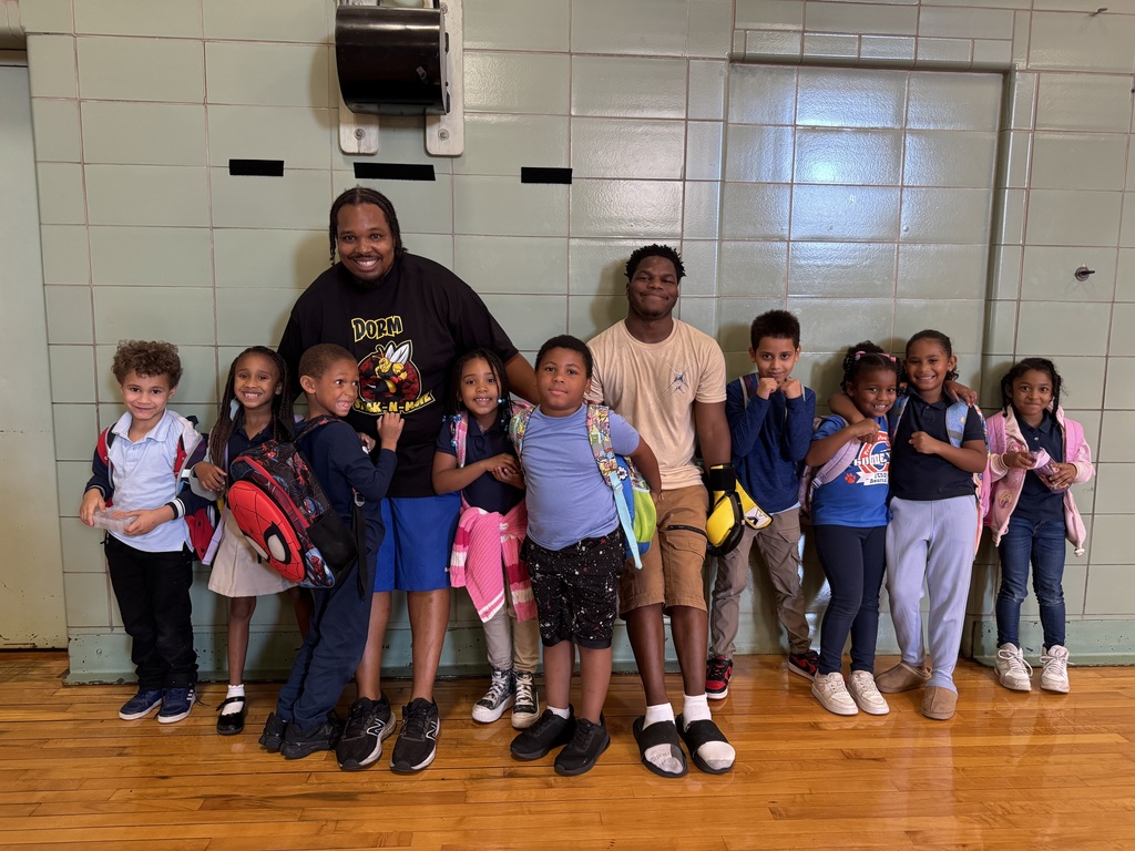Two adults and a group of young children with backpacks standing in a school gym in front of a wall.