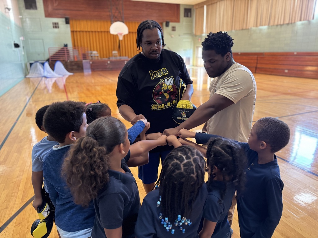 Two adults and a group of children huddling together in a school gym with their hands pointed toward the middle of a circle.
