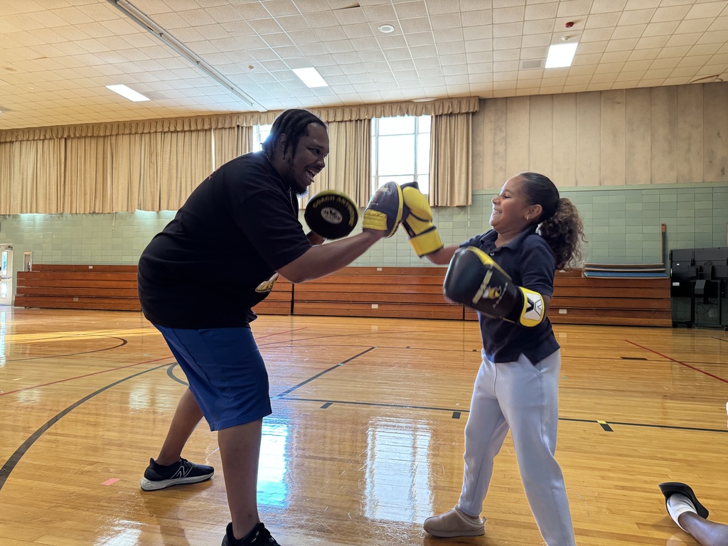 A young girl is practicing boxing with an adult in a school gym. The adult and the young girl are both wearing yellow boxing gloves.