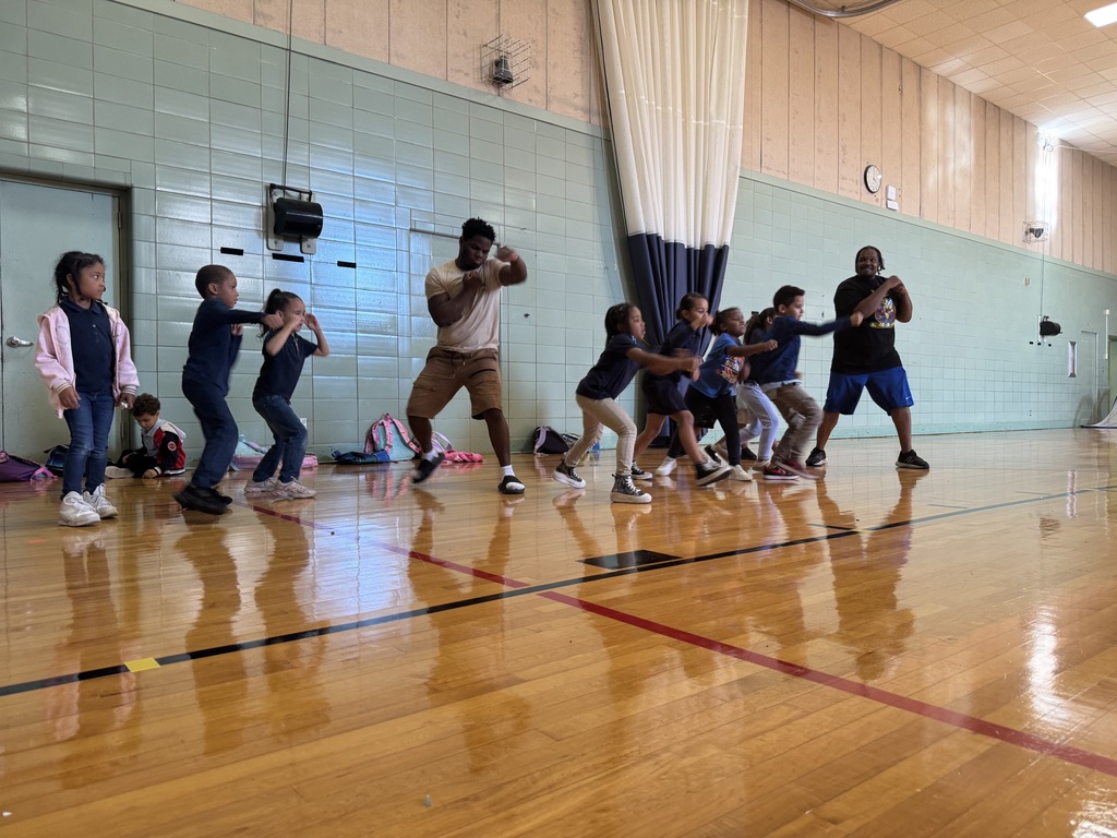 Two adults and a group of young children practicing boxing moves in a school gym in front of a wall.