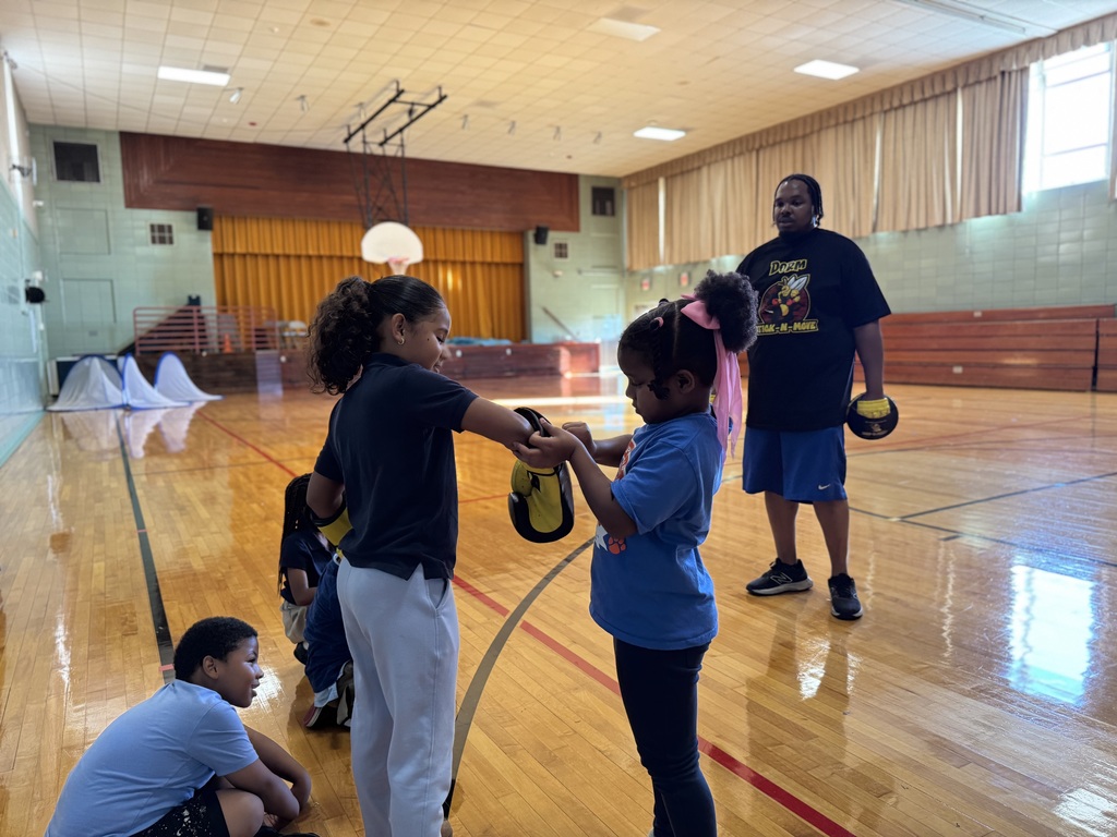 A group of children are in a school gym practicing boxing moves with yellow boxing gloves.