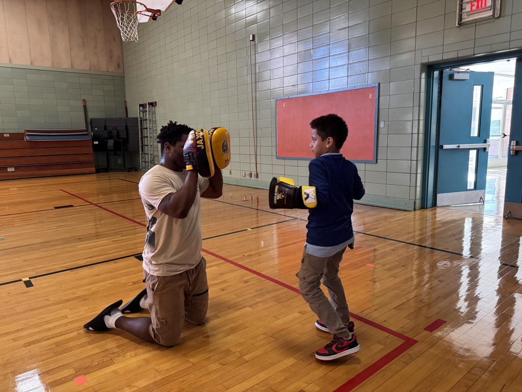 A young boy is practicing boxing with an adult in a school gym. The adult and the young boy are both wearing yellow boxing gloves.