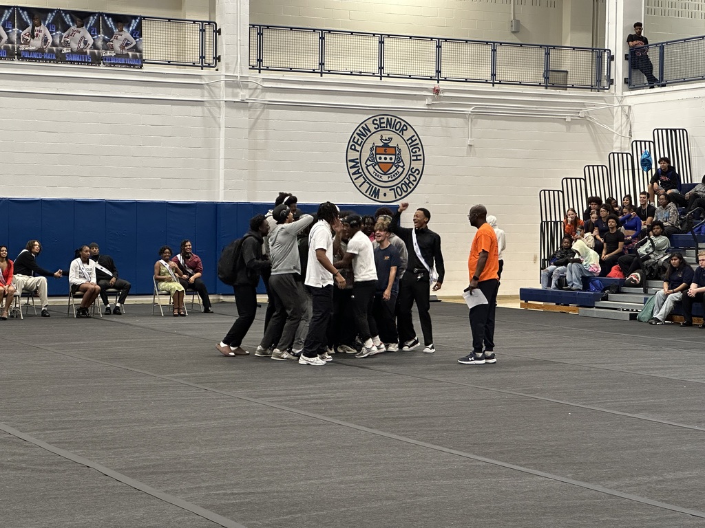 York High Boys Varsity Football Team are huddling together in a school gym on a mat as a district staff member stands near them. One of the students is holding a microphone. Other people can be seen in the background observing.