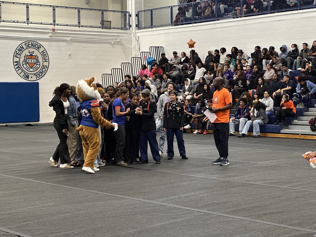 York High Girls Soccer Team are huddling together in a school gym on a mat as a district staff member and the York High mascot stands near them. One of the students is holding a microphone. Other people can be seen in the background observing.