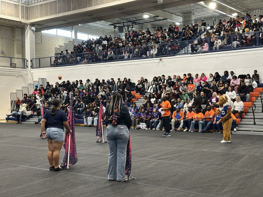 A packed school gymnasium with excited people in the bleachers. Performers in casual attire stand on the gym floor, creating a lively atmosphere.
