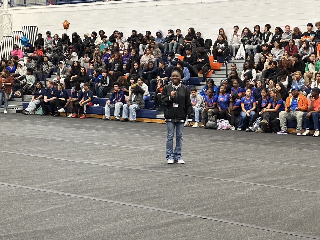 A student holding a microphone while standing on a mat in a school gym. Other people can be seen observing in the background.