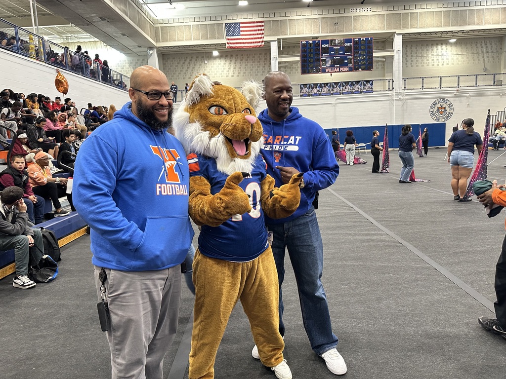 District leadership posing with the York High mascot on a mat in a school gym. Other people can be seen in the background.