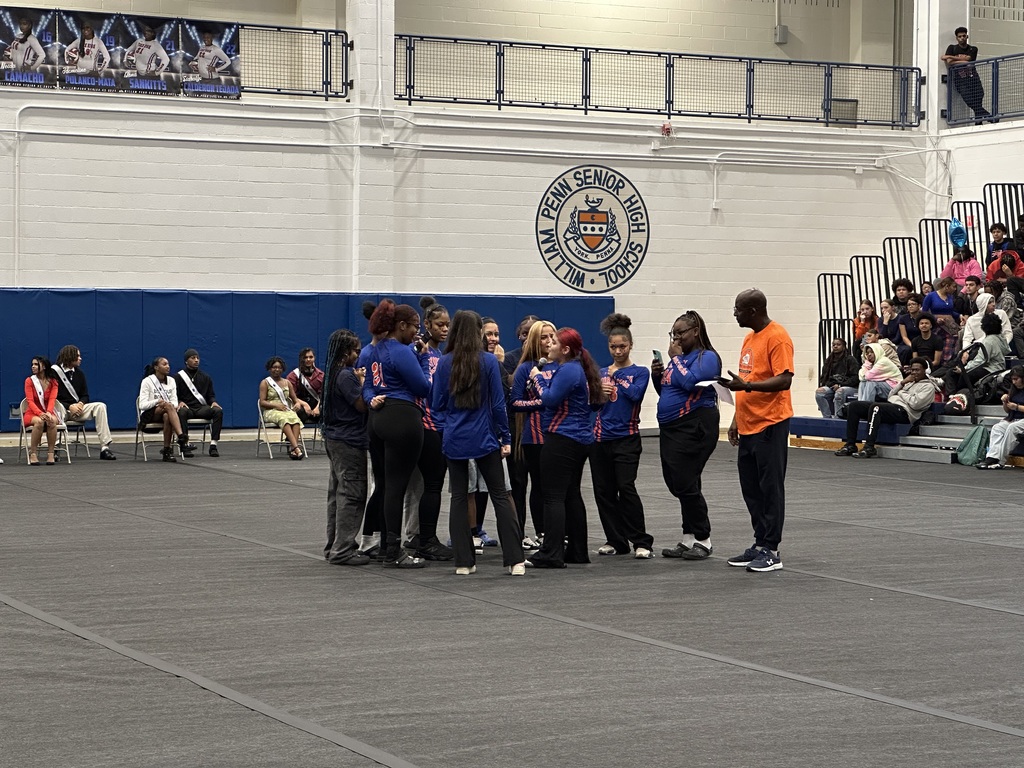 York High Girls Volleyball Team wearing blue and dark orange uniforms huddling together in a school gym on a mat as a district staff member stands near them. One of the students is holding a microphone. Other people can be seen in the background observing.