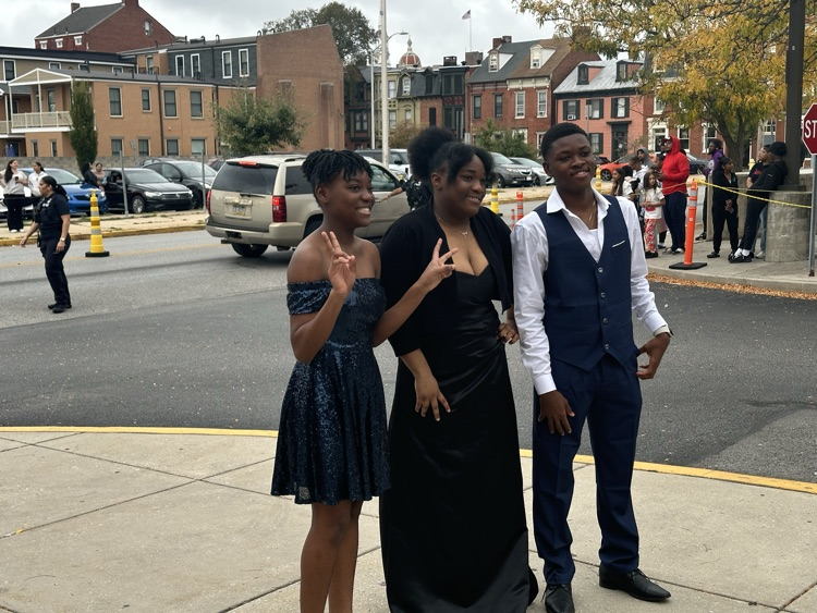 Three students are posing together outside at William Penn Senior High School’s 2025 Homecoming. Cars and other people can be seen in the background.