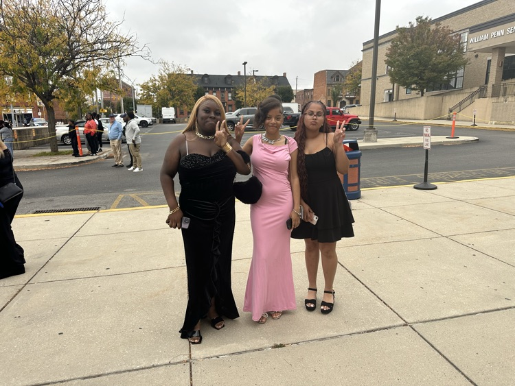 Three students are posing together outside at William Penn Senior High School’s 2025 Homecoming. Cars and other people can be seen in the background.