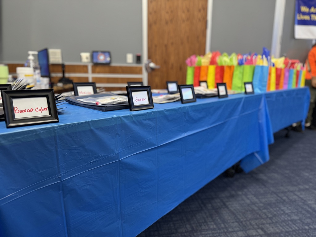 A table covered with a blue cloth displays framed name cards and documents. Brightly colored gift bags are also lined up on the table.