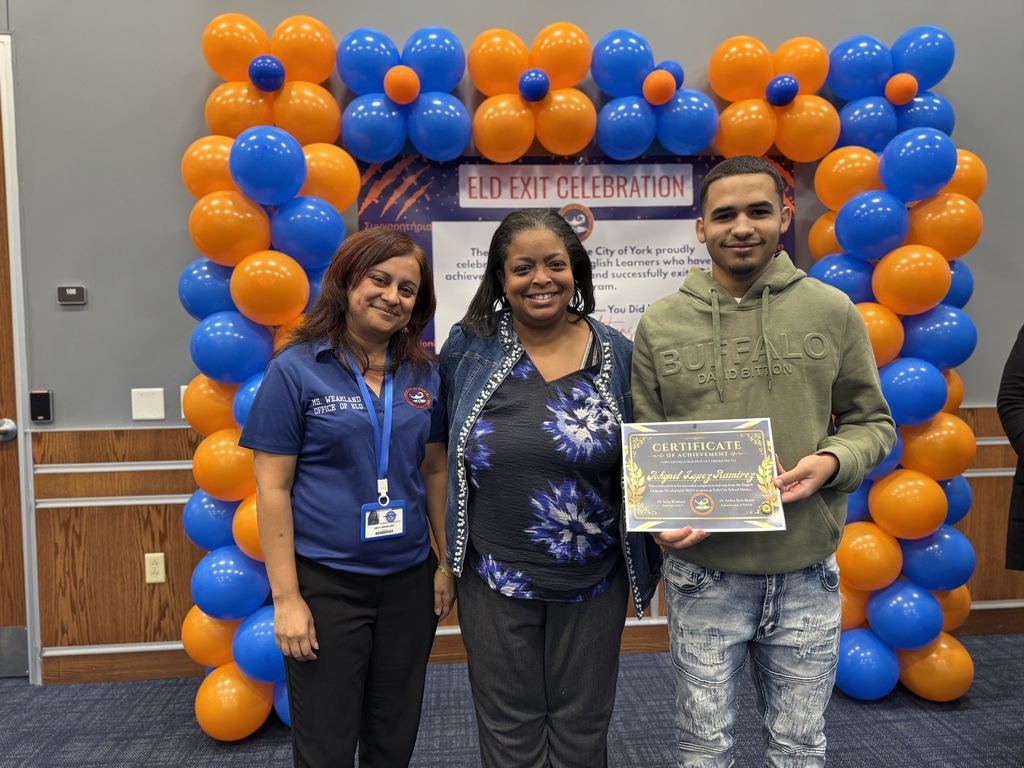 Superintendent of Schools, Dr. Andrea Berry-Brown and the district's Supervisor of ELD, Ms. Weakland posing with a student who is holding a certificate while standing in front of a banner with orange and blue balloons. The top of the banner says "ELD EXIT CELEBRATION".