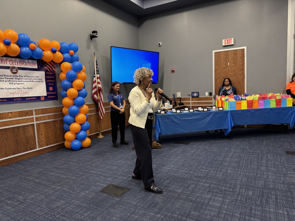Representative Carol Hill-Evans is speaking into a microphone at a celebratory event. Behind her are blue and orange balloons, a poster, a U.S. flag, and tables with colorful gift bags. Superintendent of Schools, Dr. Andrea Berry-Brown and the district's Supervisor of ELD, Ms. Weakland are standing nearby. 