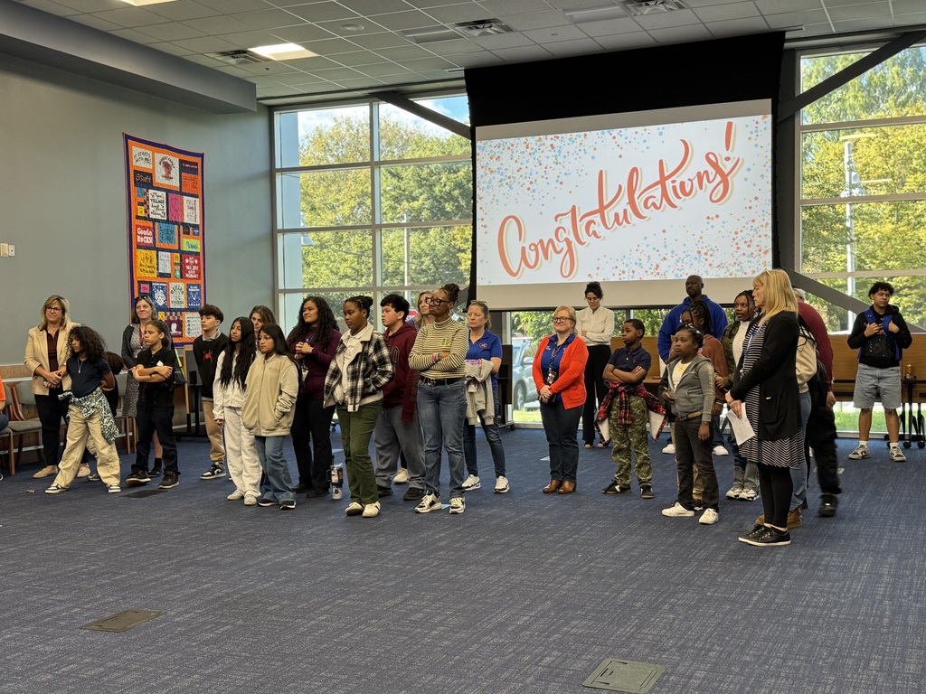 A diverse group of students and teachers standing attentively in a classroom, presenting a unified and engaged atmosphere. A screen is displaying "Congratulations!" with confetti graphics.