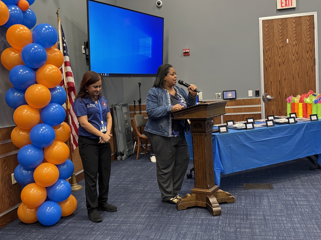 Superintendent of Schools, Dr. Andrea Berry-Brown is speaking at a podium in a room that is decorated with orange and blue balloons. Behind her is a U.S. flag. The district's Supervisor of ELD, Ms. Weakland is standing nearby Dr. Andrea Berry-Brown. There are awards on a table that is covered with a blue tablecloth.