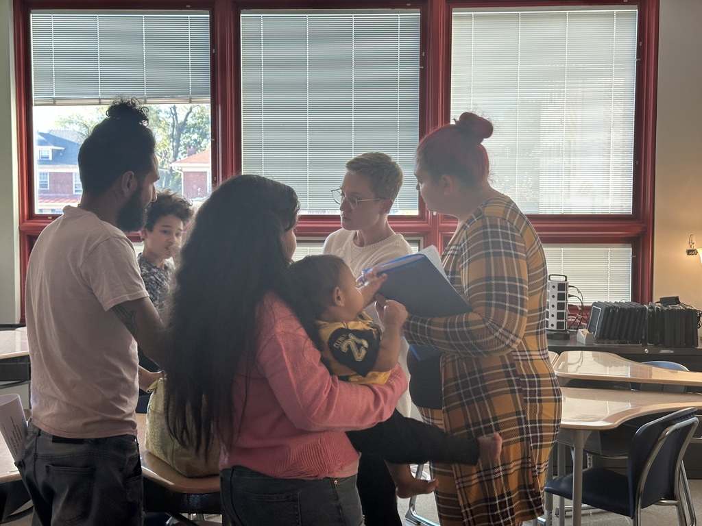 A group of people are gathered in a classroom, engaging in a discussion. A girl is holding a child, while one of the adults is holding a notepad. 