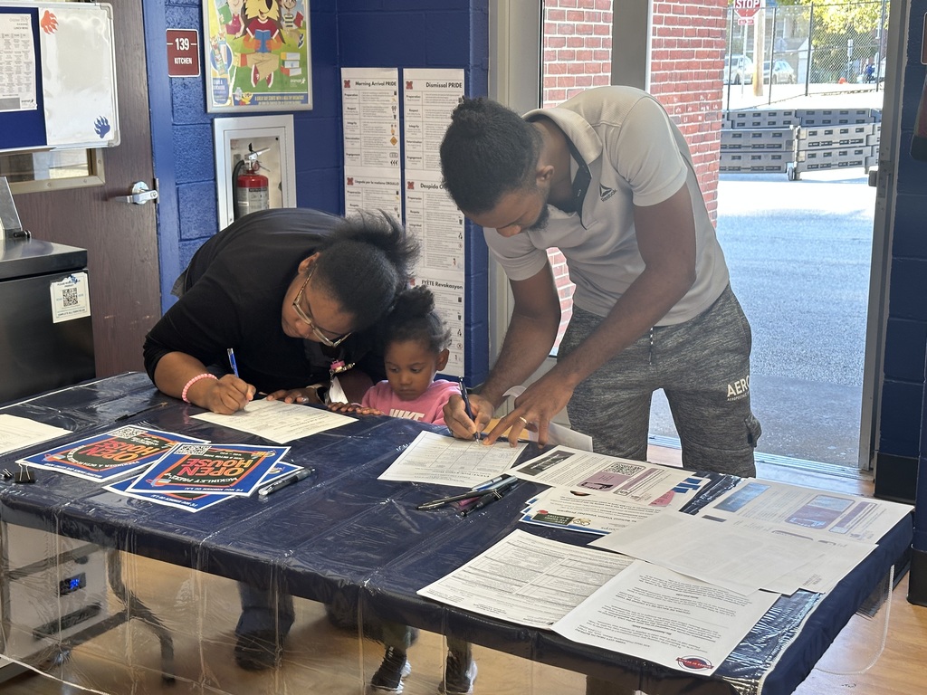 Two adults are filling out forms at a table that is covered with papers and flyers. A small child can be seen looking down at the table.