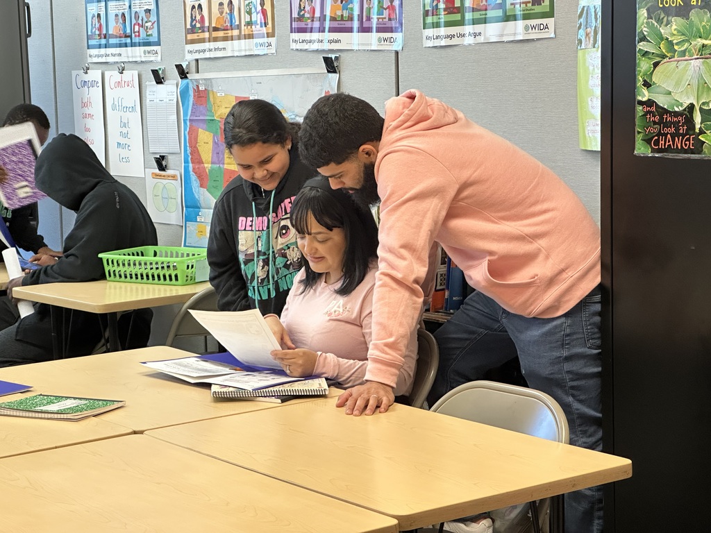 Two adults and one student are reviewing materials at a table in a classroom, with educational posters visible on the walls.