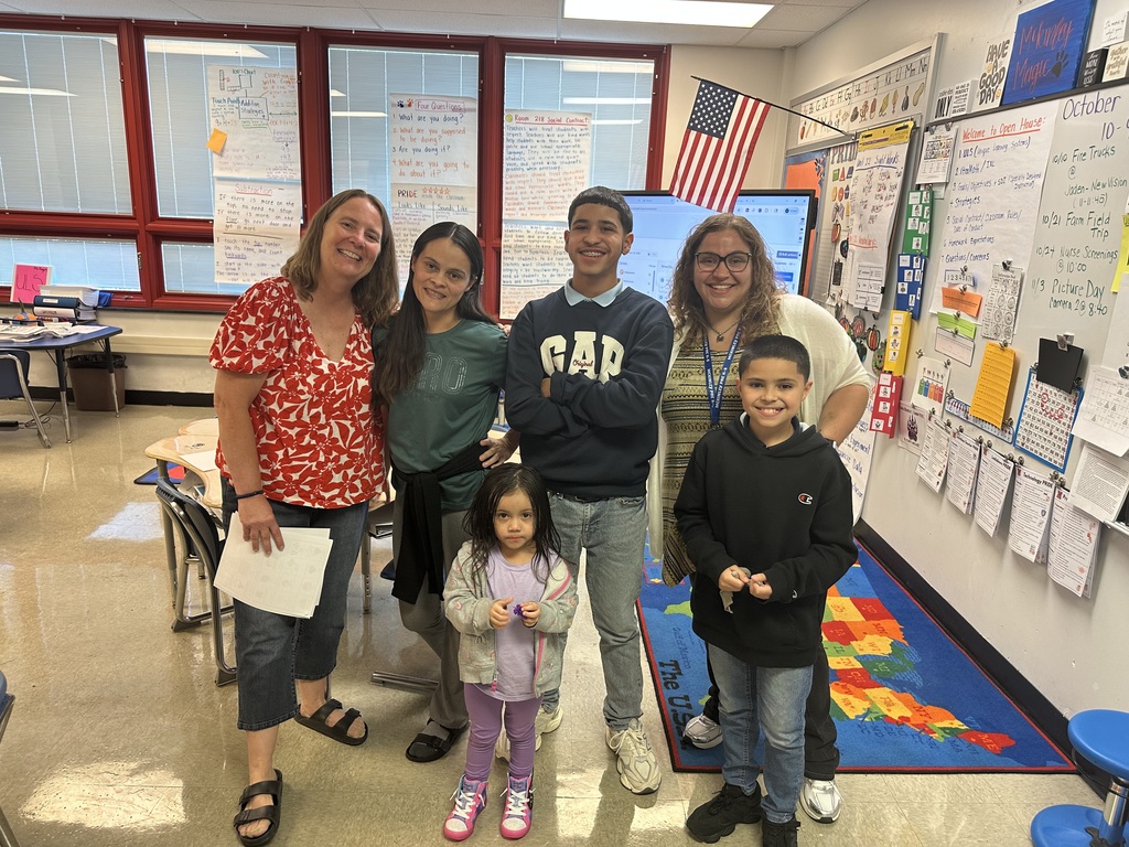 A group of six people, including children and adults, are posing together in a classroom with educational materials displayed on the walls.